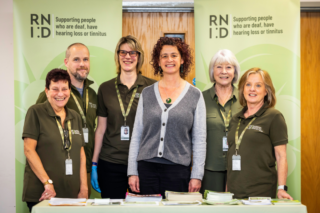RNID Near You volunteers and RNID CEO, Harriet Oppenheimer, stand in front of green RNID banners.