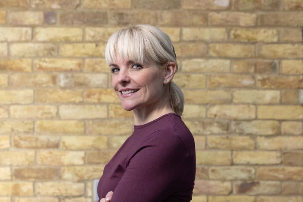 Charlotte stands in front of a brick wall with her arms crossed. She has blonde hair and wears a cochlear implant. 