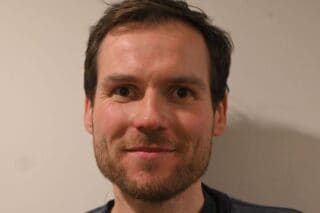 A headshot of a smiling main with brown hair and facial hair.