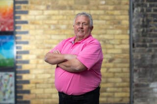 RNID storyteller, Dougie, wears a pink t-shirt and stands in front of a brick wall with his arms folded.