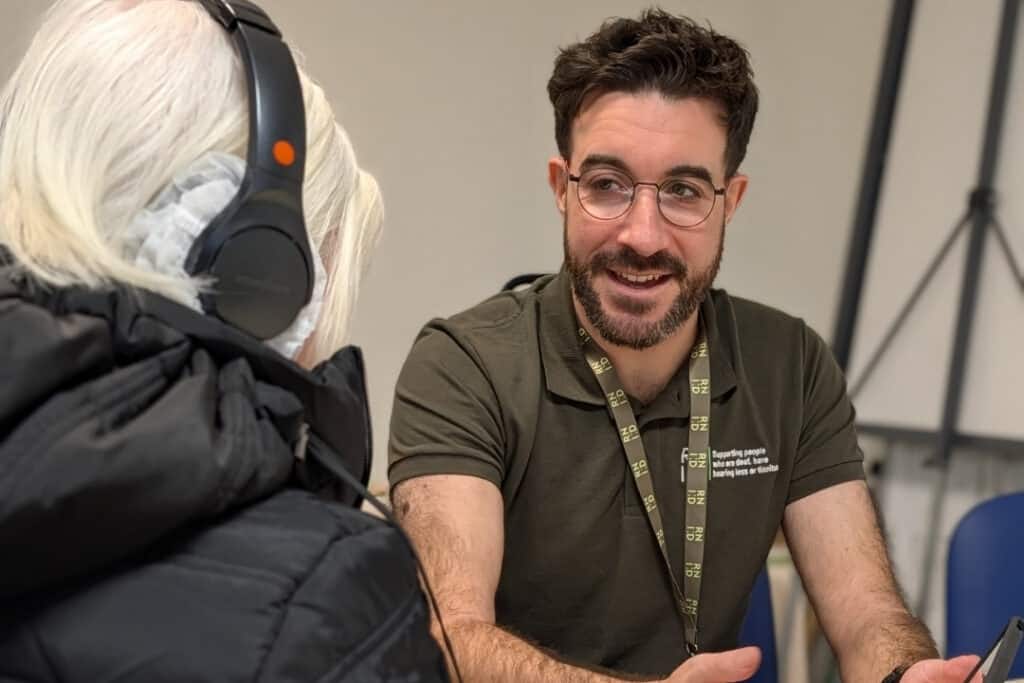 A man with brown hair and beard wears a green RNID Near You t-shirt and helps someone take a hearing check.
