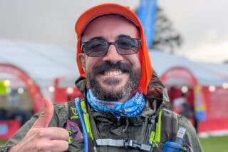 A man wears glasses and an orange cap with a running vest holding water. He holds his thumb up to the camera and is smiling.