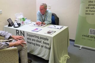 RNID Near You volunteer, Bruce, sits behind a desk helping someone with their hearing aids.