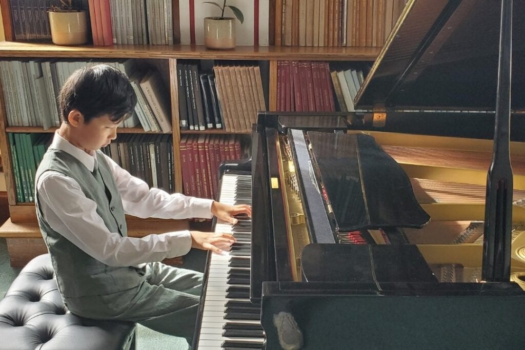 A boy wears a waistcoat, shirt and trousers. He sits on a stool, playing the piano.