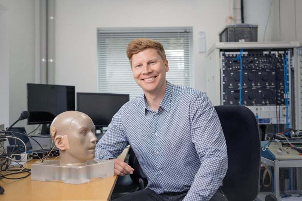 A man with blonde hair wearing a blue checked shirt sits at a desk with scientific equipment around him.