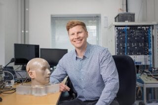A man with blonde hair wearing a blue checked shirt sits at a desk with scientific equipment around him.