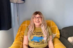 A person with long blonde hair wearing glasses sits in a yellow armchair and is smiling.