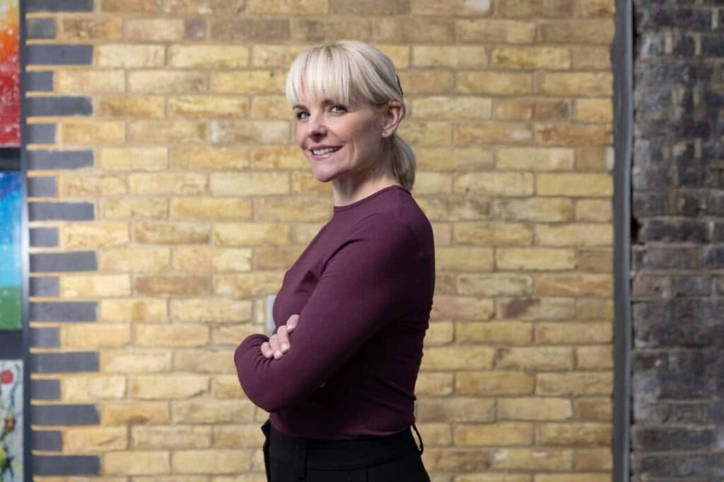 A woman with blonde hair stands in front of a brick wall with arms folded and smiles. She wears a cochlear implant.