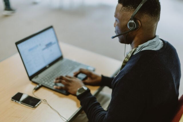 A man sits at a desk using a laptop wearing a headset.