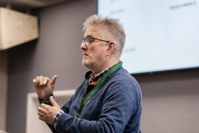 A man wearing a blue shirt and green lanyard signs to an audience.