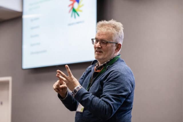 A man stands in front of a large screen presentation and uses BSL to communicate with the audience.