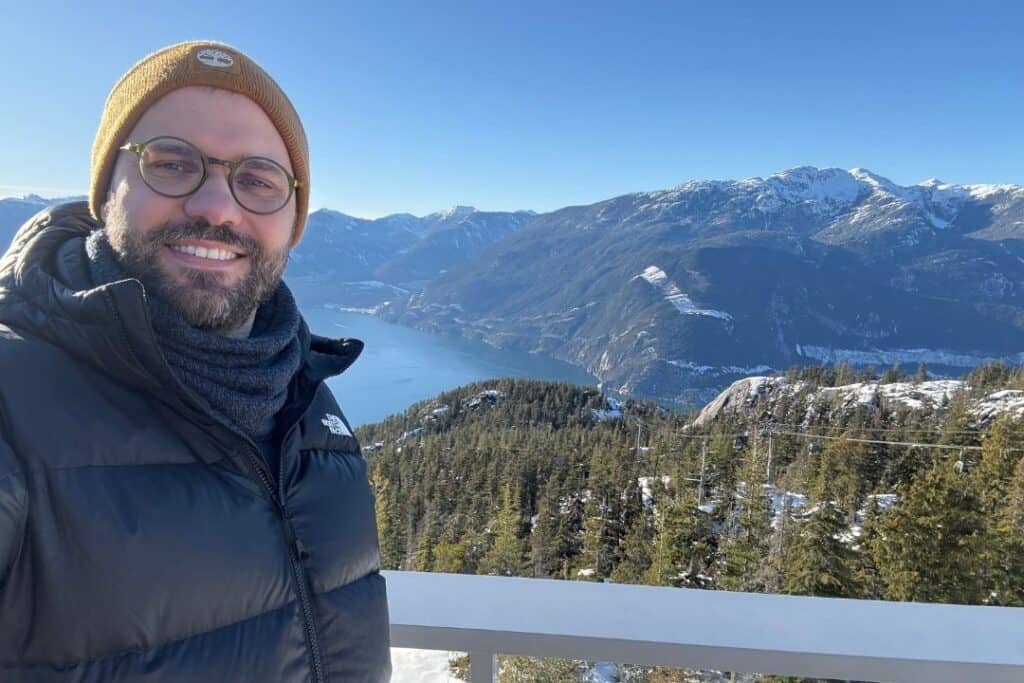 RNID storyteller, Tomasz, stands on a balcony overlooking a forest. He is smiling, and wearing a yellow beanie hat and glasses.