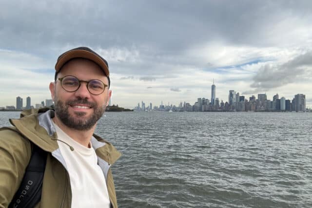 RNID storyteller, Tomasz, stands by the sea. He wears a cap and glasses.