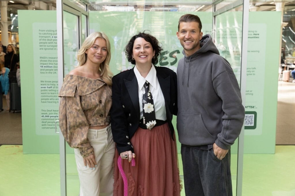 Celebrities Tasha Ghouri, Sam Baines, and Jamie Clarke stand in front of a clear box during Deaf Awareness Week.