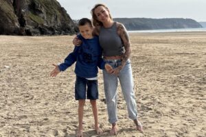 A teeange boy with short hair wears a jumper and denim shorts. He stands beside his mum who wears jeans and a grey vest. They are on a sandy beach, holding hands and smiling.