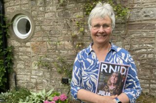 RNID storyteller, Mel, stands in front of a brick wall and holds an RNID members Magazine.