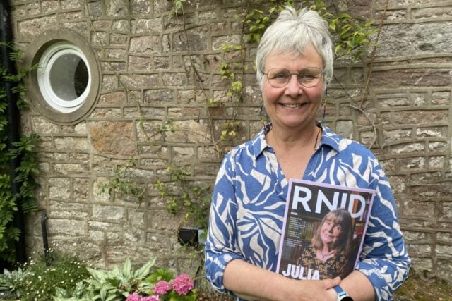 RNID storyteller, Mel, stands in front of a brick wall and holds an RNID members Magazine.