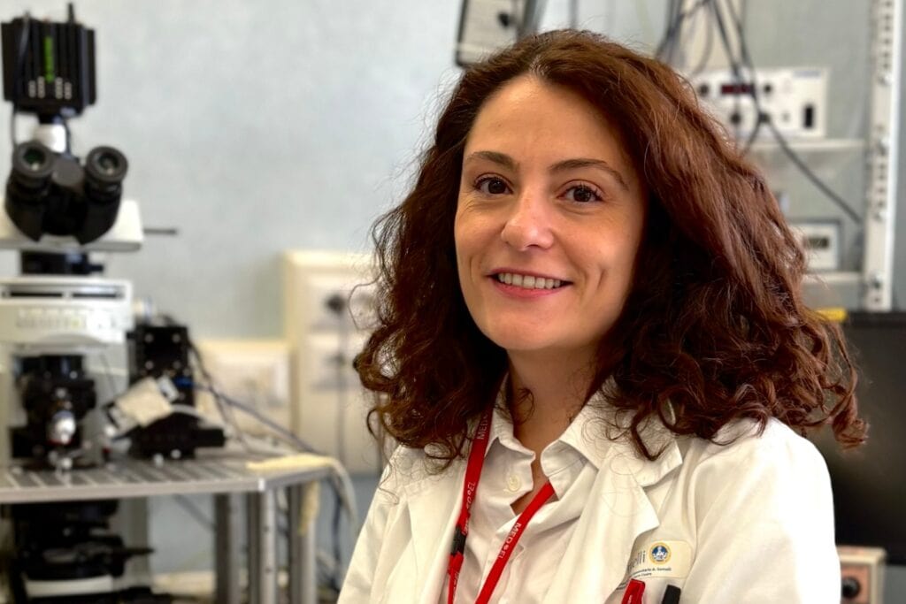 A female researcher with shoulder length curly brown hair wears a lab coat and sits in a laboratory.