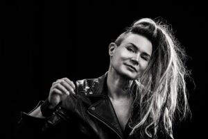 Jenni from Deafmetal wears a black leather jacket and has her hair to one side with her head tilted.